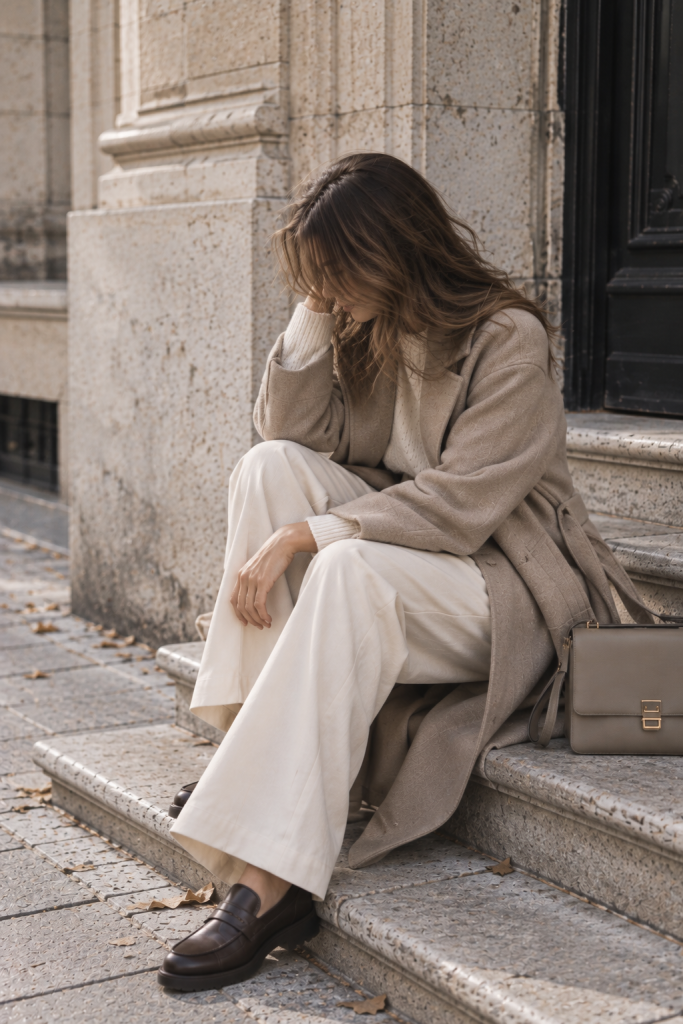 Sitzender Herbstlook mit taupefarbenem Mantel, hellem Strickpullover, Stoffhose und dunklen Loafern