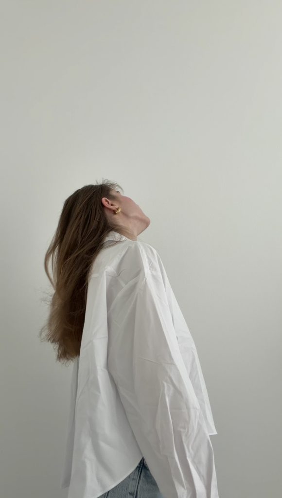 Frau in weißer Oversize-Bluse mit langen braunen Haaren vor heller Wand