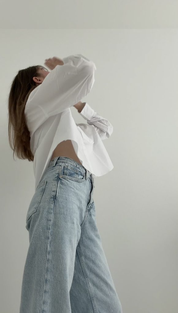 Frau in weißem Oversize-Hemd und heller Jeans vor minimalistischer Wand – moderner Ausdruck lässiger französischer Eleganz.