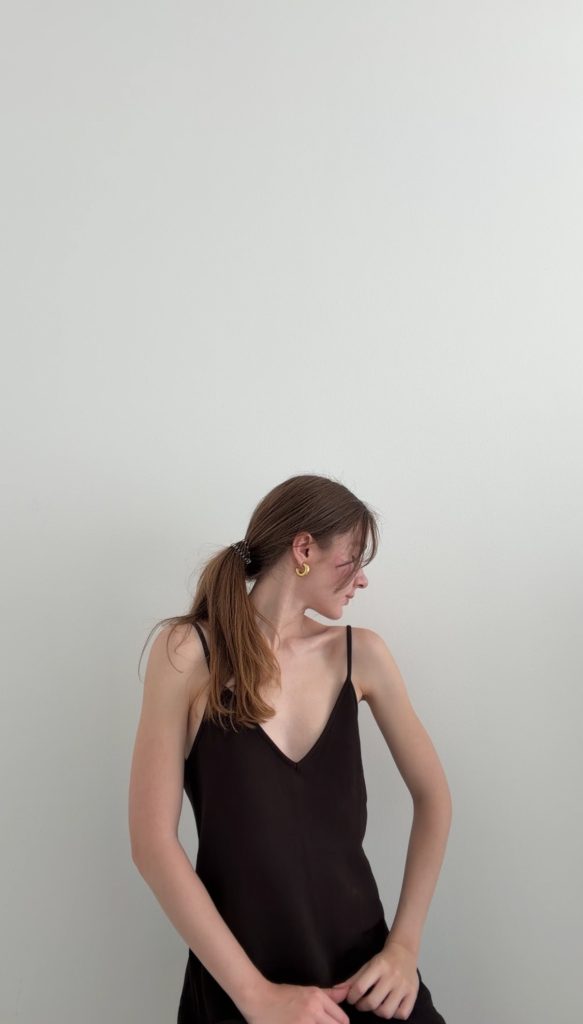 Sitzende Pose in einem dunkelbraunen Kleid mit V-Ausschnitt und schmalen Trägern vor einer schlichten weißen Wand.