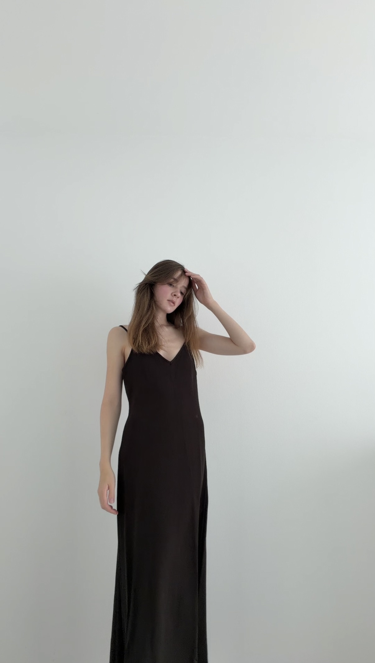 Outfit Proportionen in einer minimalistischen Ganzkörperaufnahme mit langem dunkelbraunem Trägerkleid vor weißem Hintergrund.