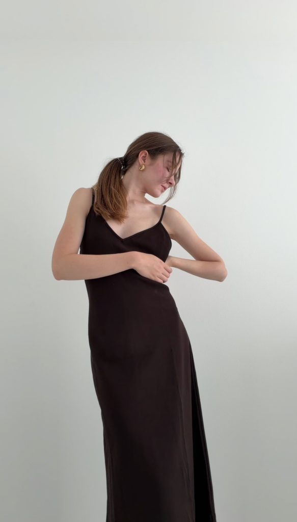 Dunkelbraunes Maxikleid mit fließender Linienführung, seitlich angewinkelten Armen und leicht geneigtem Oberkörper.